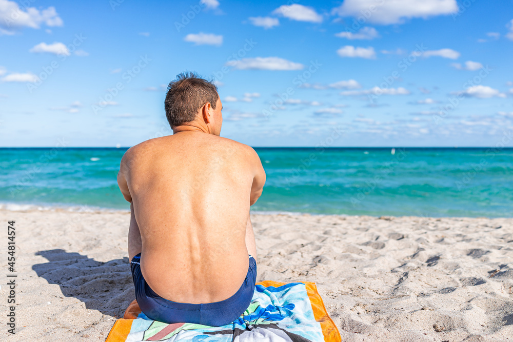 Turquoise vibrant colorful water and back of tan man sitting on towel looking at sand shore in Sunny Isles Beach, North Miami, Florida during sunny day in winter Turquoise vibrant colorful water and back of tan man sitting on towel looking at sand shore in Sunny Isles Beach, North Miami, Florida during sunny day in winter