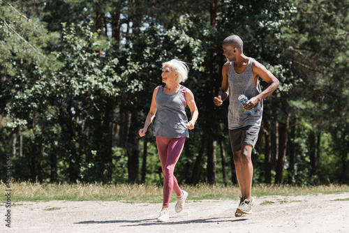Smiling senior sportswoman jogging near african american husband with sports bottle in forest.