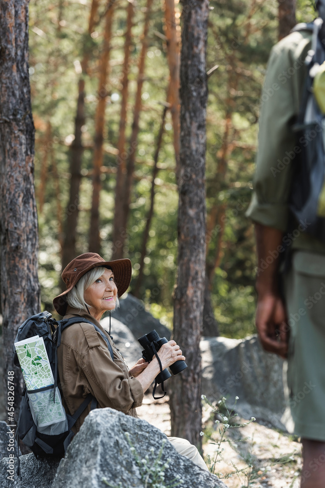 Smiling senior woman holding binoculars near blurred african american husband in forest.