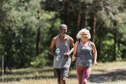 Smiling elderly sportswoman running near african american husband with sports bottle in forest.
