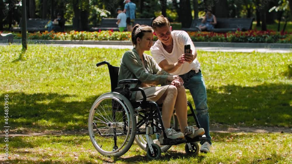 Woman in a wheelchair and her friend spending time at the summer park and taking a selfie together Woman in a wheelchair and her friend spending time at the summer park and taking a selfie together