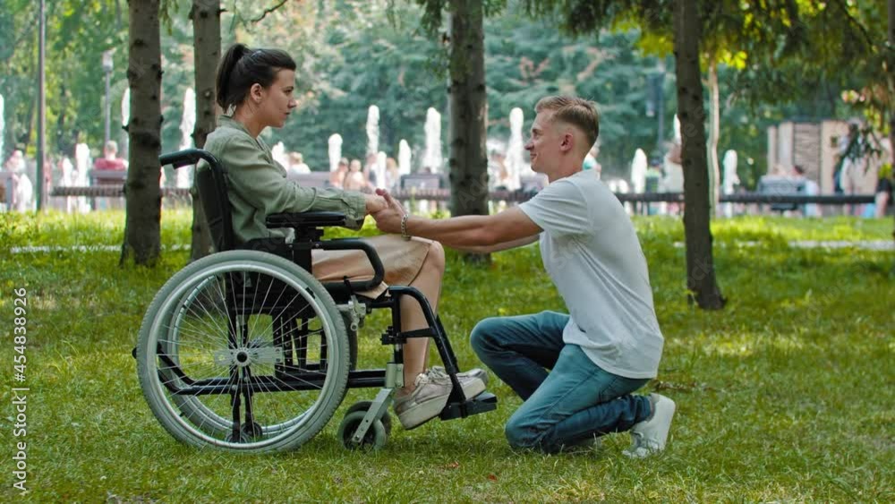 Alone woman sitting in a wheelchair in the park and her boyfriend comes up to her and holds her hands standing on the knees Alone woman sitting in a wheelchair in the park and her boyfriend comes up to her and holds her hands standing on the knees