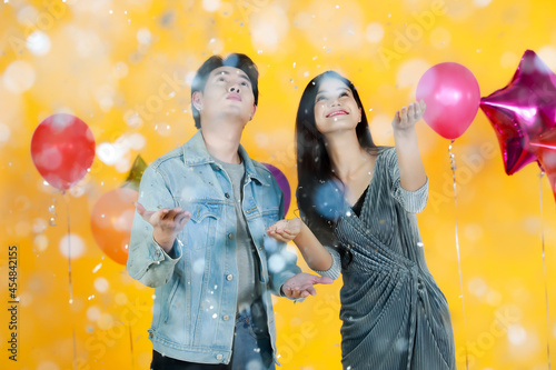 Studio shot of Asian happy young male and female lover couple model in fashionable wears play andi fun withpaper confetti in celebration party with balloons on yellow background