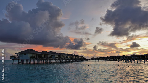 A colorful sunset over the Indian Ocean. There are purple, scarlet, and golden clouds in the blue sky. Above the aquamarine water - two rows of water villas. Maldives © Вера 