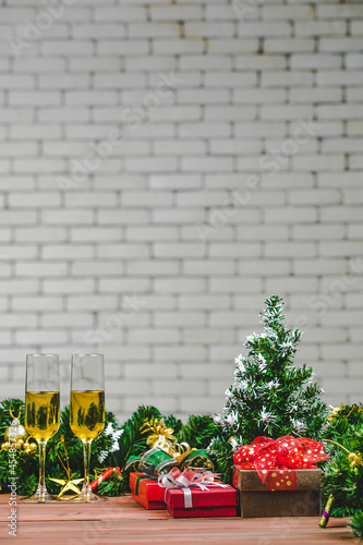 Champagen two glasses next to red and gold gift boxes, a artificial small pine tree, christmas ornaments, prepare for celebrate put wood board, white brick wall blur background photo