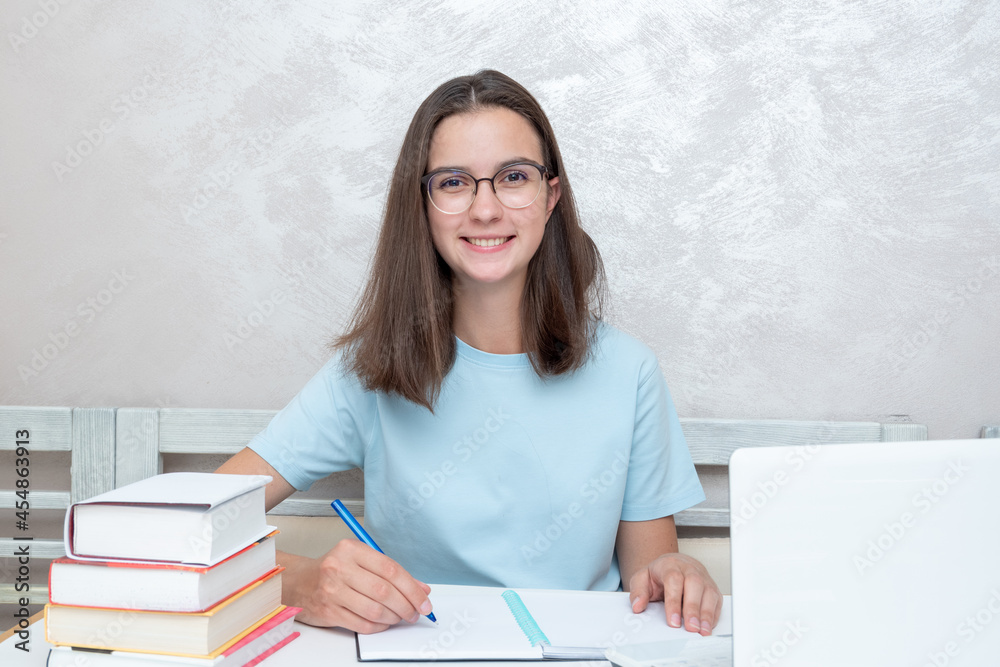 A smiling teenager student sitting at a table with books is writing an assignment in a notebook. A smiling teenager student sitting at a table with books is writing an assignment in a notebook.