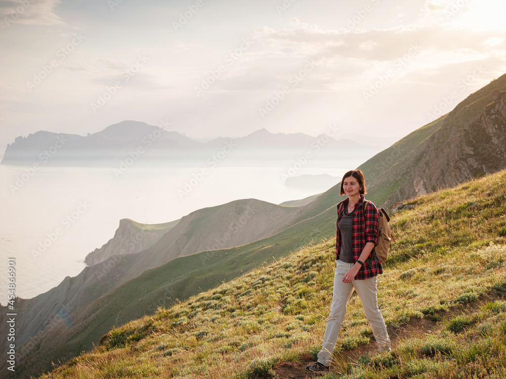 A young Asian woman with a backpack hiking in the summer climbing the mountain at sunset . Mountain and coastal travel, freedom and an active lifestyle
