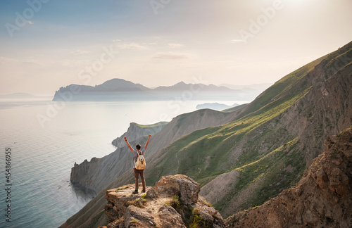 Young man travels alone on the backdrop of the mountains standing on top of a mountain with raised hands, the lifestyle concept of traveling outdoors. © YURII Seleznov