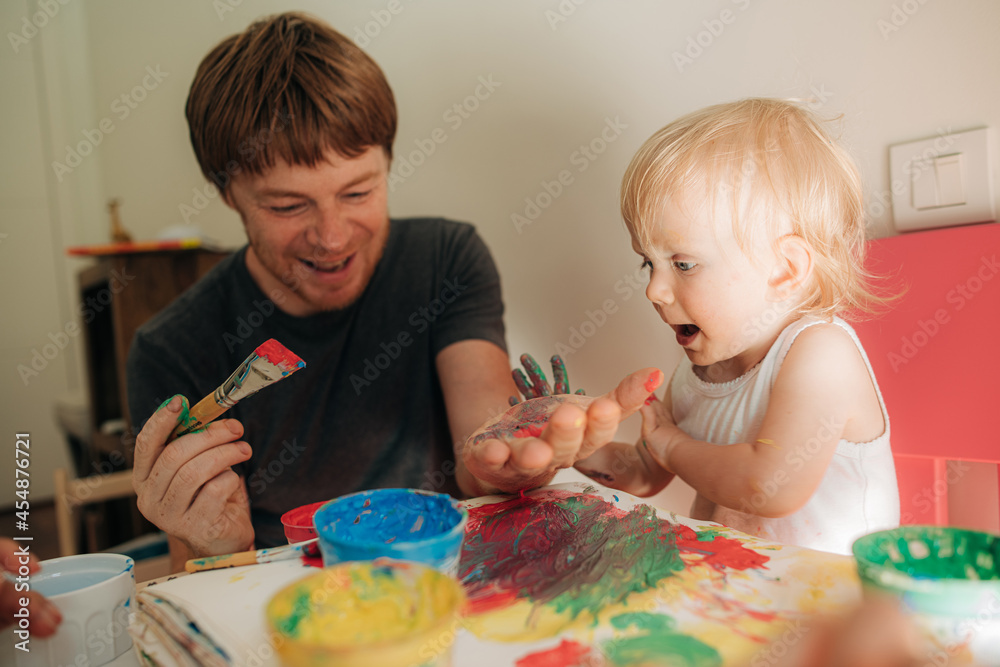 Fototapeta premium Joyful father and daughter playing with paints. Man painting with his little child at home. Creativity concept