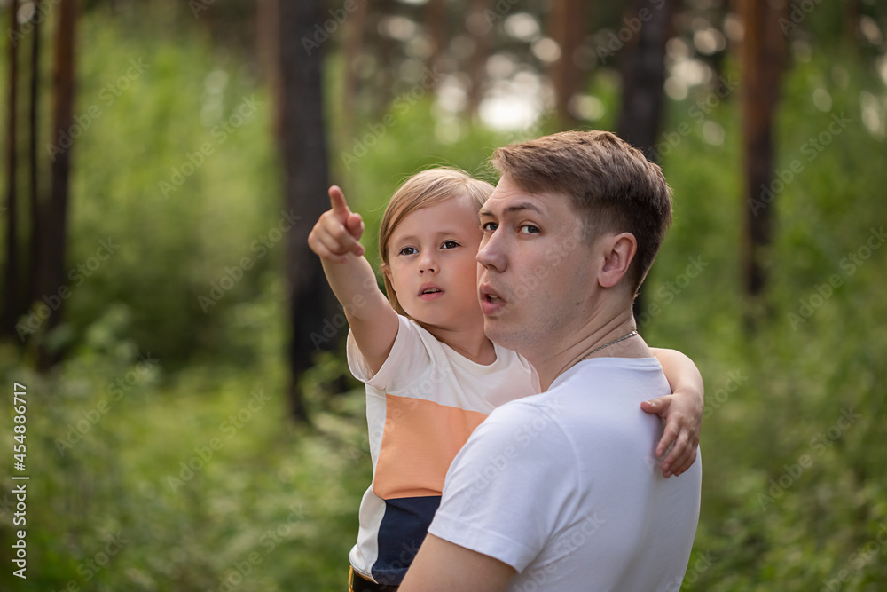 Caucasian daughter in daddy's arms points a finger to the side in the forest. Caucasian daughter in daddy's arms points a finger to the side in the forest.