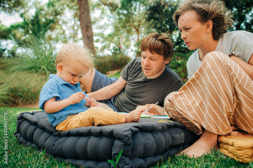 Portrait of family painting with child outdoors. Confident parents sitting on mattress with toddler daughter in park. Parenthood and creativity concept