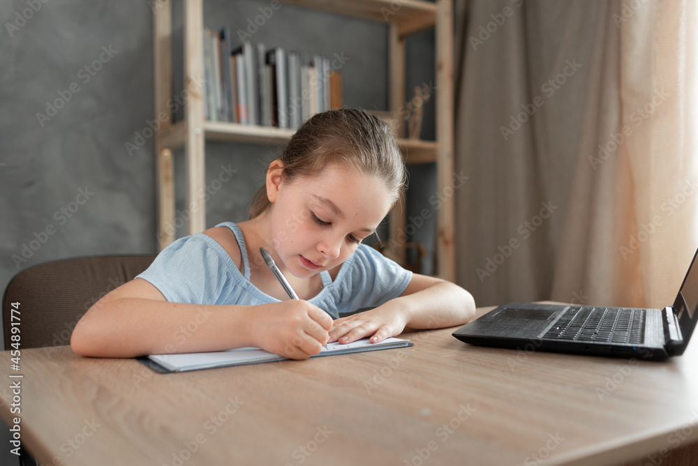 Cute teenage schoolgirl doing homework using laptop at home. The child writes down the task in a notebook. Cute teenage schoolgirl doing homework using laptop at home. The child writes down the task in a notebook.