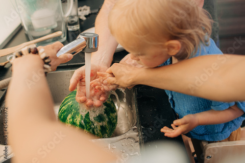 Little girl washing hands with mother and father at kitchen. Close-up of family with little child washing watermelon in sink. Family at home, childhood, hygiene concept
