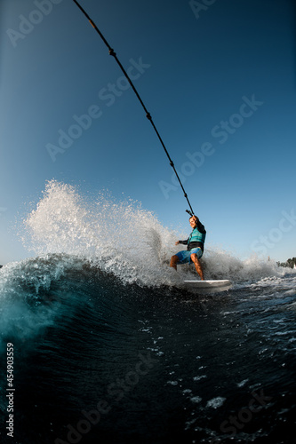 man riding on the wakesurf holding rope of motorboat on the background of blue sky