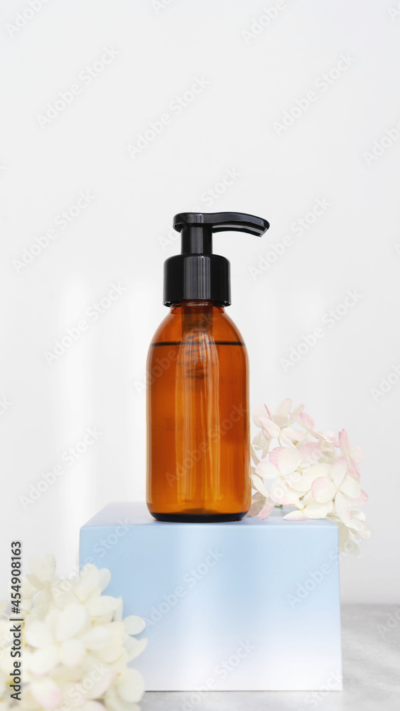  brown glass cosmetic bottle with dispenser  for soap or shampoo on blue podium with flowers on white background with sunlight. Beauty product Vertical