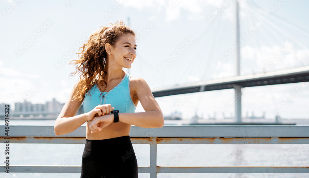 Obraz premium Uses a smart watch to measure the pulse on the hands. A woman does sports on the street. Fitness and a healthy lifestyle. The trainer does exercises for stretching the body.