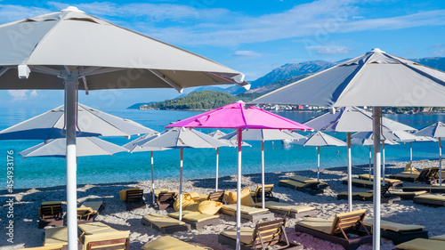 One pink open beach umbrella among the rest of grays. Large beach umbrellas over sun loungers on pebble beach.