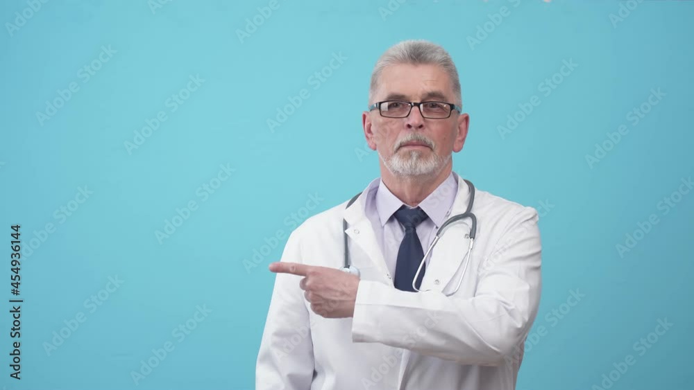 Serious man doctor points finger to side and looking into camera. Adult doctor in a medical uniform points finger to the side, standing in the studio with a blue background. Serious man doctor points finger to side and looking into camera. Adult doctor in a medical uniform points finger to the side, standing in the studio with a blue background.