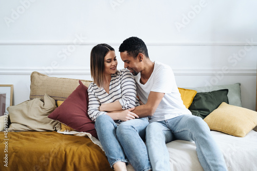 Portrait of happy young African Caucasian couple hug sitting on couch at home, smiling mixed race husband and wife spend time together, embrace relaxing on sofa. Healthy relationships concept