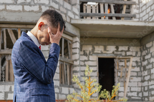 distressed young man in business suit stands in front of destroyed unfinished house, the inability to pay for housing, construction or mortgage crisis