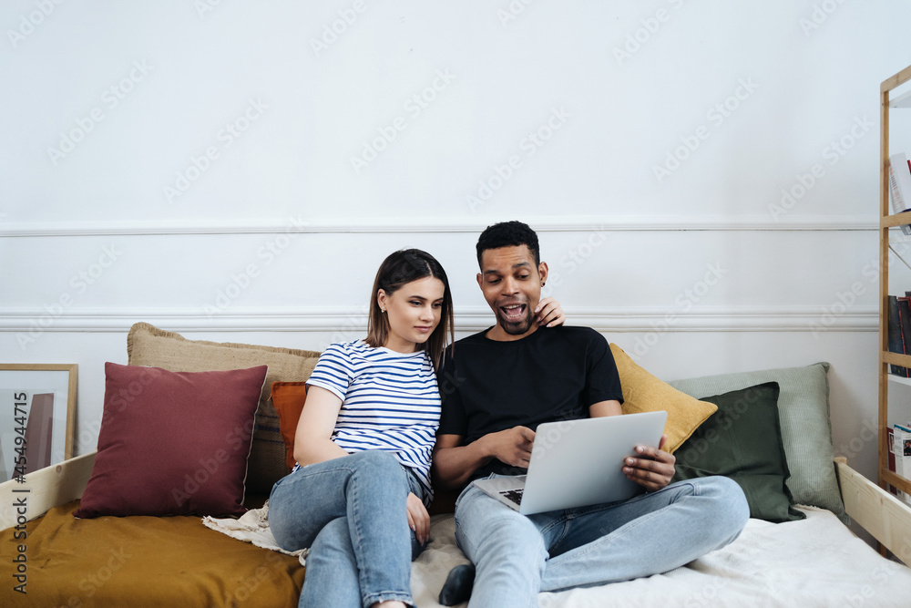 Happy young mixed race couple enjoying time at home using computer sitting on sofa, black man and white woman having fun online shopping or surfing web relaxing on couch Happy young mixed race couple enjoying time at home using computer sitting on sofa, black man and white woman having fun online shopping or surfing web relaxing on couch