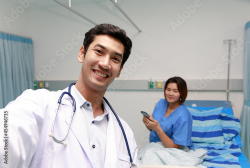 Young and friendly smart doctor smile and looking at camera during taking selfie photo with female patient on bed in hospital room