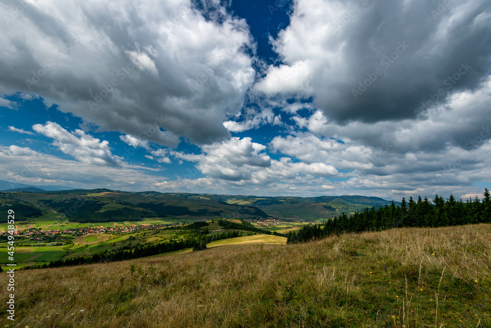 Wide angle view from the top of the mountain, small hungarian village in the valley, blue sky with white clouds in Transylvania, Romania.