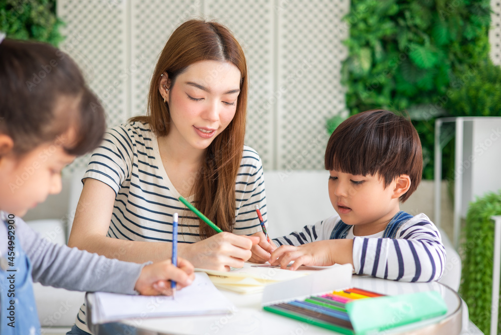 happy loving family. pretty young mother reading a book and drawing to her daughter and son.Mother teach Asian preschool student do homework by reawing by a color. happy loving family. pretty young mother reading a book and drawing to her daughter and son.Mother teach Asian preschool student do homework by reawing by a color.