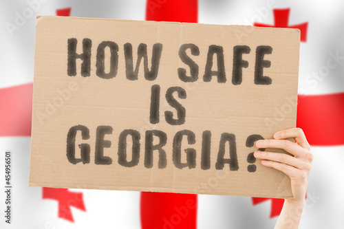 The question " How safe is Georgia? " on a banner in men's hand with blurred Georgian flag on the background. Safety. Street. Outdoor. Dangerous. Security. Attack. Criminal. Criminality
