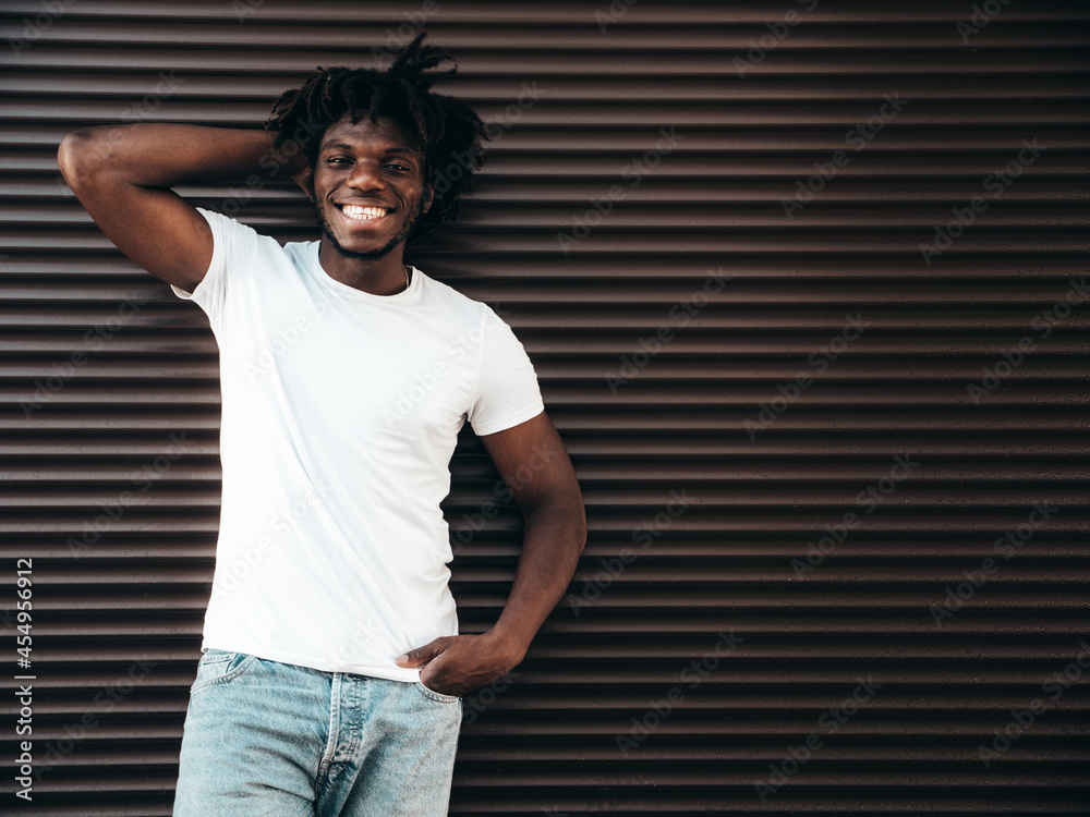 Portrait of handsome hipster  smiling African man dressed in  white summer t-shirt and jeans. Fashion male with dreadlocks hairstyle  posing near roller shutter wall in the street Stock Photo | Adobe