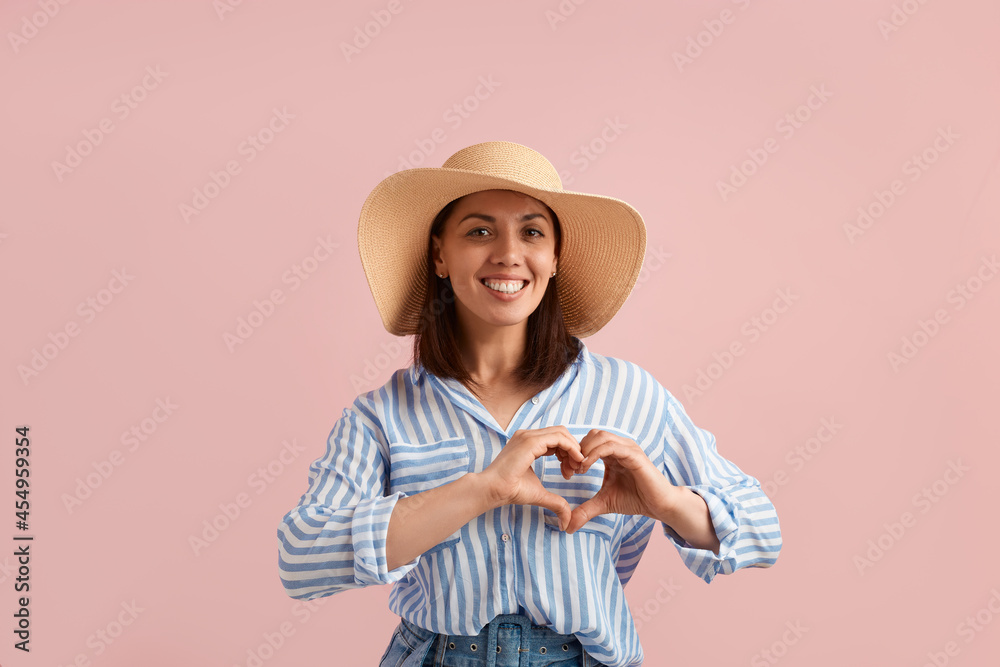 Attractive smiling brunette cowgirl confesses in love, makes heart gesture, shows feelings, has happy expression, wears straw hat, striped shirt, jeans, on pink background. Summer emotions concept. Attractive smiling brunette cowgirl confesses in love, makes heart gesture, shows feelings, has happy expression, wears straw hat, striped shirt, jeans, on pink background. Summer emotions concept.