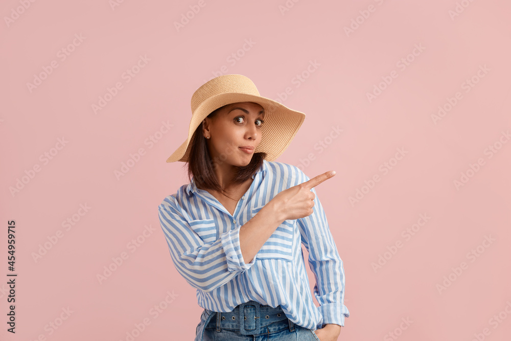 Impressed woman points out at blank copy space with index finger, recommends product, has eyes wide open, wears straw hat, striped shirt, jeans with belt, on pink background. Summer emotions concept. Impressed woman points out at blank copy space with index finger, recommends product, has eyes wide open, wears straw hat, striped shirt, jeans with belt, on pink background. Summer emotions concept.