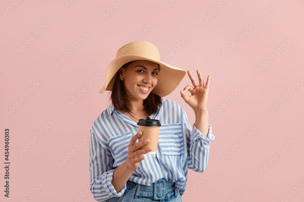 Approved. Smiling happy woman with dark hair is showing ok gesture, holds takeaway hot morning coffee, wears straw hat, striped shirt, jeans with belt, on pink background. Summer emotions concept. Approved. Smiling happy woman with dark hair is showing ok gesture, holds takeaway hot morning coffee, wears straw hat, striped shirt, jeans with belt, on pink background. Summer emotions concept.