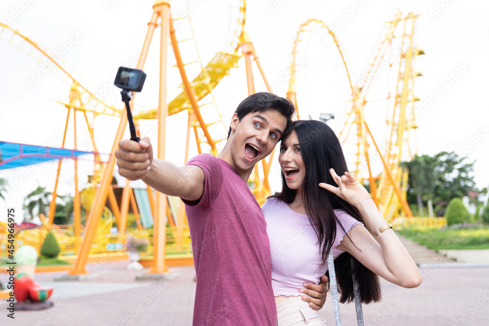 Obraz premium Cheerful caucasian young couple having fun while talking selfie at the amusement theme park on Roller coaster background. Attractive happy couple at the theme park. couple, people, holiday concept