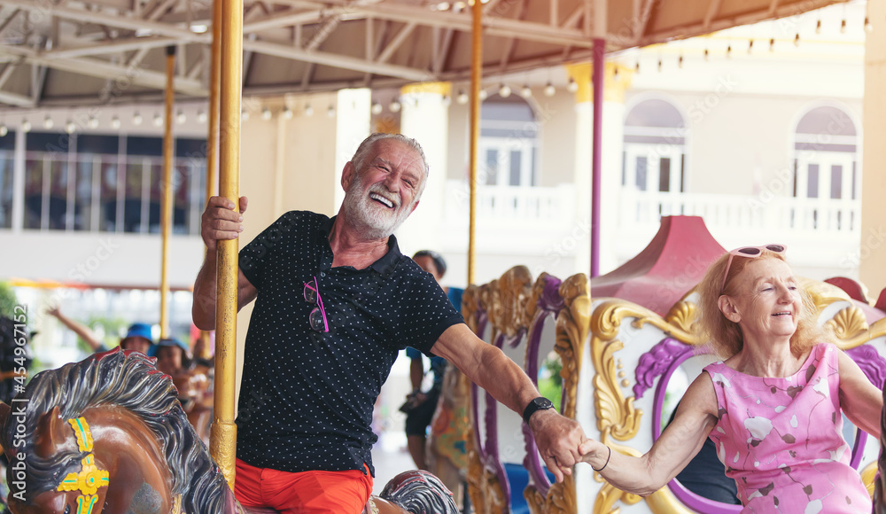 Happy cheerful smiling senior couple holding hands together and playing on horse carousel ride at amusement park. Elderly couple having fun at the theme park. people, tourist and holiday concept