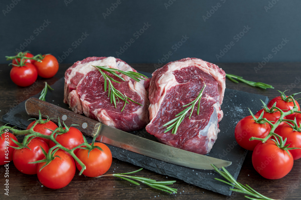 Close-up view of raw beef meat (Chuck steak) lying on dark table next to red fresh tomatoes, kitchen knife and small green rosemary branches. Fresh organic food theme.