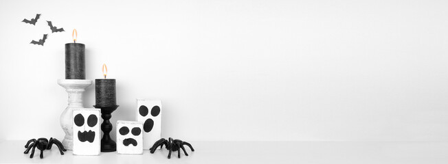 Halloween shelf display with rustic wood ghost decor, black candles and spiders against a white wall with bats. White shelf. Banner with copy space.