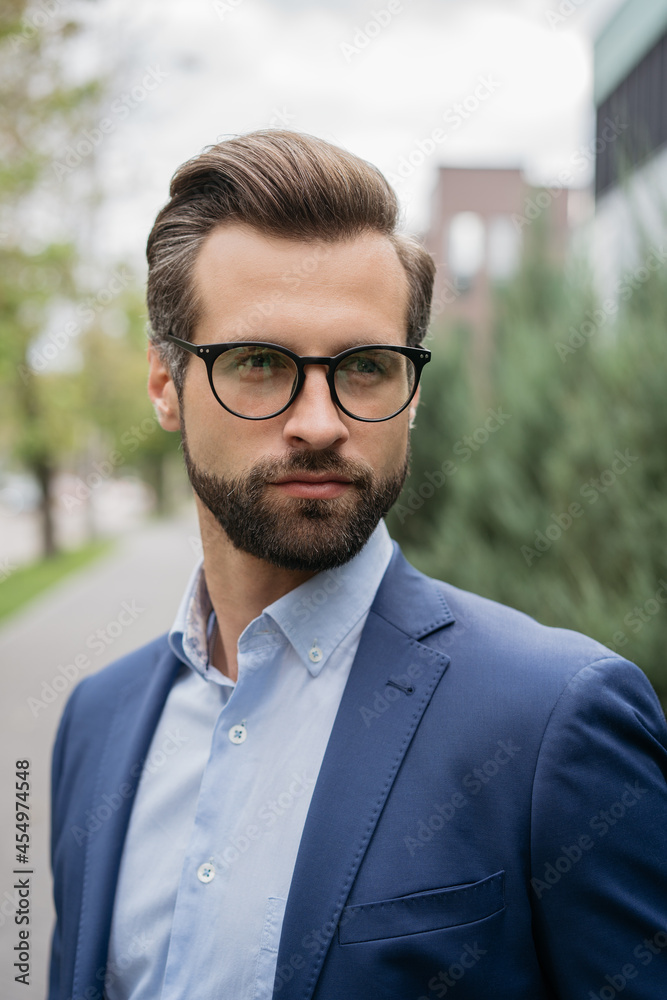 Handsome bearded man with stylish hair wearing business suit and glasses looking away on the street. Closeup portrait of young confident banker standing outdoors. Career concept 