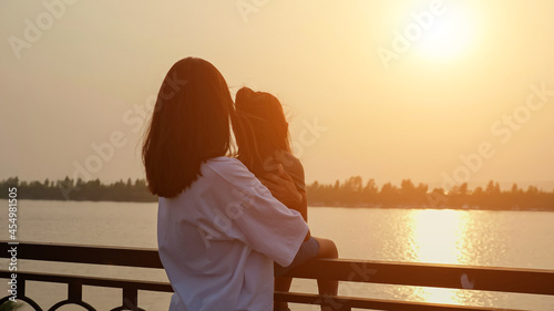 Φωτογραφία Mother holds little girl sitting on railing to enjoy bright sunset on contempora