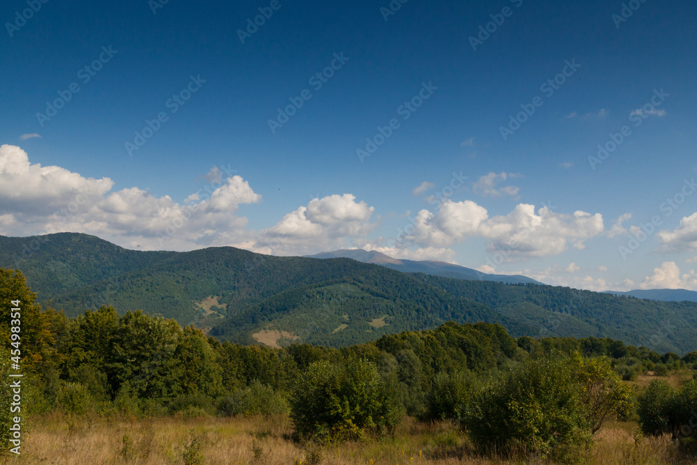 Mountain meadow in the light of the morning sun. Rural summer landscape with a valley in the fog behind the forest on a grassy hill. Fluffy clouds in the bright blue sky. Nature freshness concept. Car