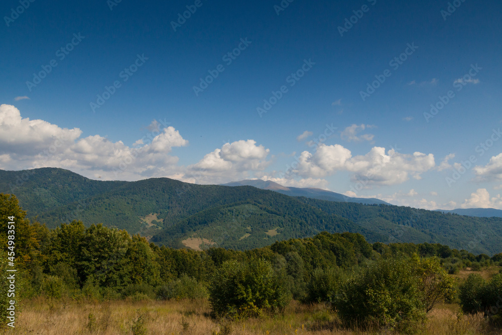 Mountain meadow in the light of the morning sun. Rural summer landscape with a valley in the fog behind the forest on a grassy hill. Fluffy clouds in the bright blue sky. Nature freshness concept. Car Mountain meadow in the light of the morning sun. Rural summer landscape with a valley in the fog behind the forest on a grassy hill. Fluffy clouds in the bright blue sky. Nature freshness concept. Car
