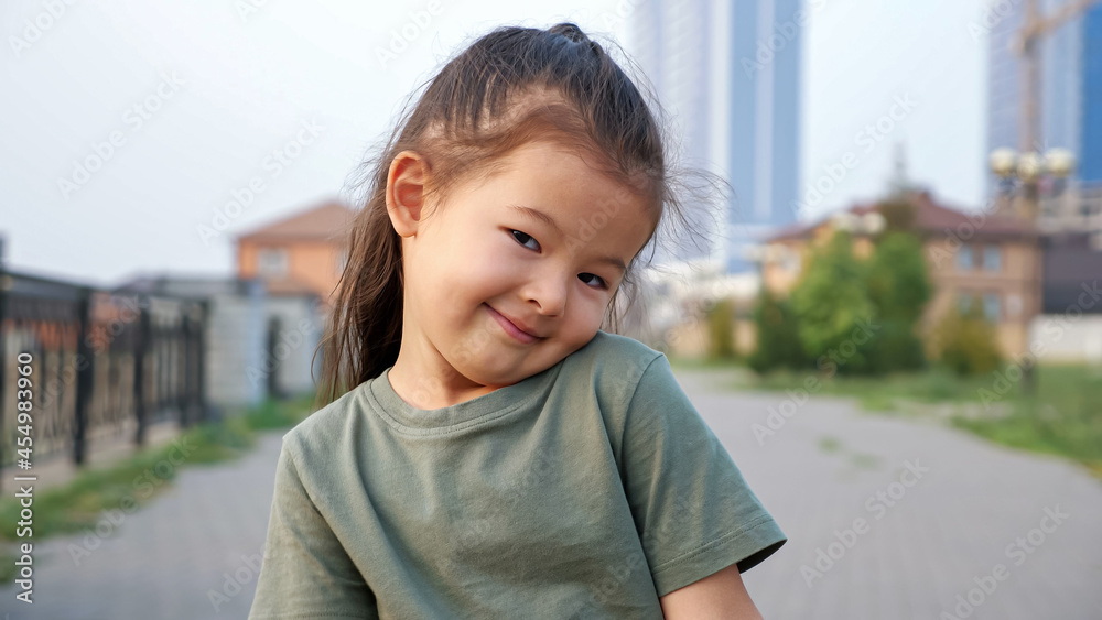 Adorable happy little Asian girl in green t-shirt poses for camera standing on city waterfront against buildings in summer evening close view Adorable happy little Asian girl in green t-shirt poses for camera standing on city waterfront against buildings in summer evening close view