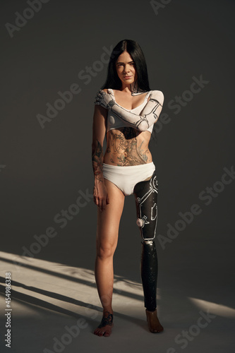 Full length view of the tattooed female with cyborg pattern on her body posing at the studio on dark background. People and robot concept