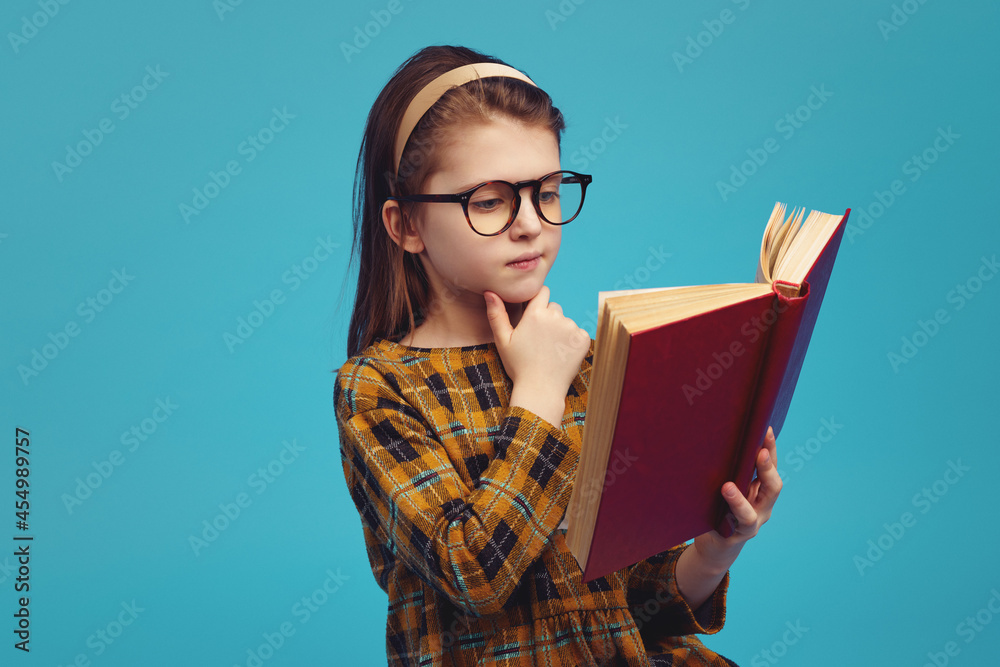 Adorable schoolgirl wearing checkered dress and eyeglasses standing against blue color background and reading book, doing homework Adorable schoolgirl wearing checkered dress and eyeglasses standing against blue color background and reading book, doing homework