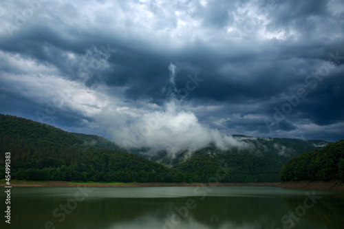 Impressive landscape of green coniferous mountains above lake against the background of stormy gray clouds, rainy weather, Ukraine, Carpathians