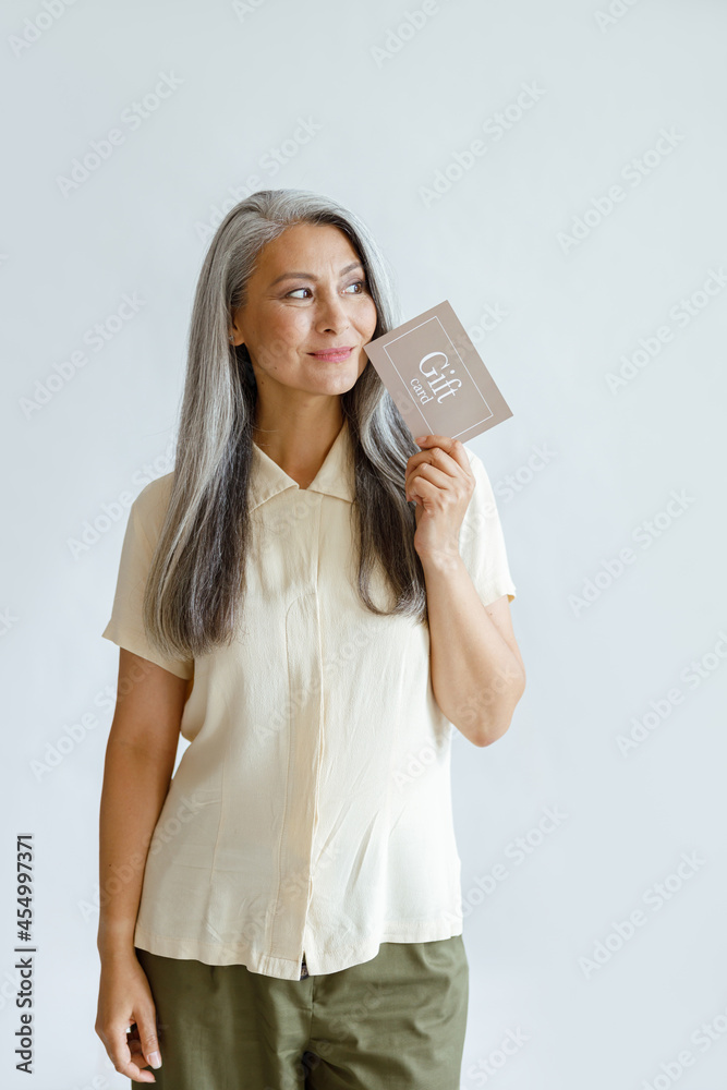 Naklejka premium Thoughtful hoary haired Asian lady customer holds gift card standing on light grey background in studio. Shopping certificate