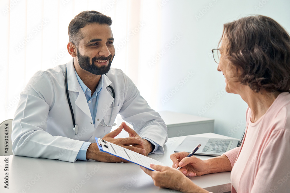 Happy Indian male doctor consulting senior old patient filling form at  consultation. Friendly professional physician talking to mature woman  signing medical paper during appointment visit in clinic. Stock-Foto