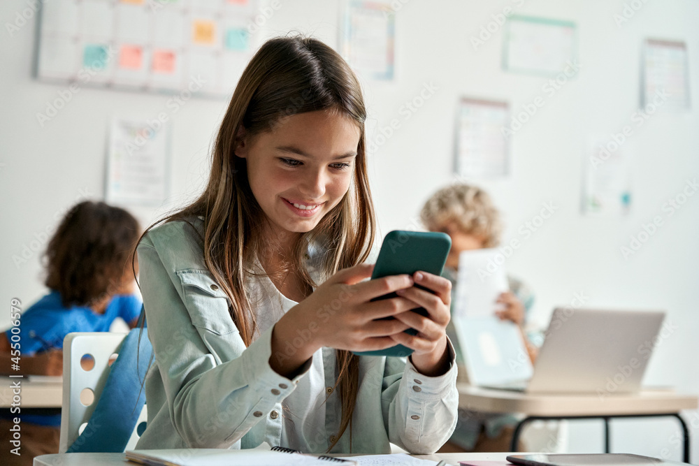 Happy latin hispanic kid girl school student using smartphone in classroom. Preteen child holding mobile cell phone having fun with apps playing games and checking social media at school during break. Happy latin hispanic kid girl school student using smartphone in classroom. Preteen child holding mobile cell phone having fun with apps playing games and checking social media at school during break.