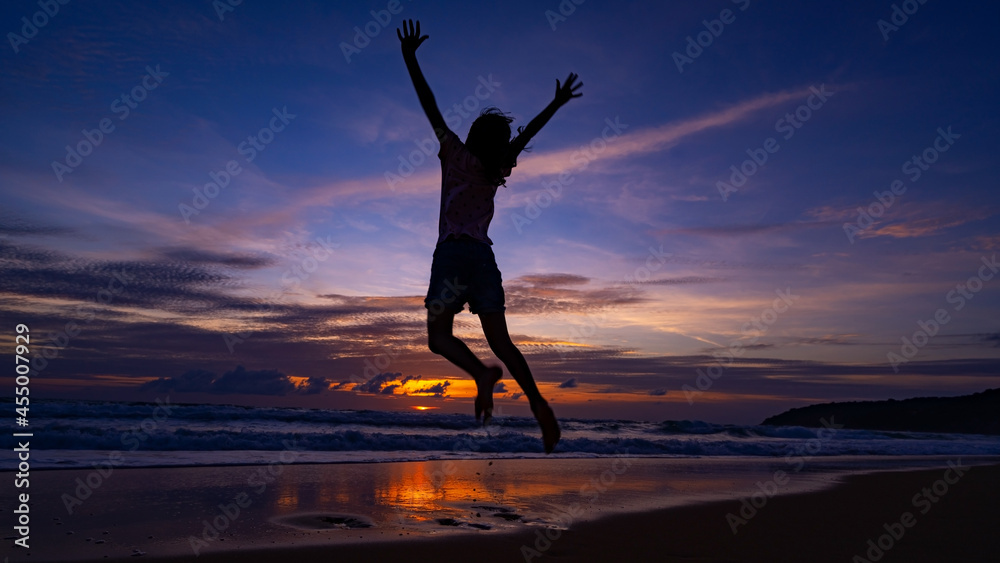 Silhouette of happy kid girl jumping on the beach at sunset or sunrise sky over sea.