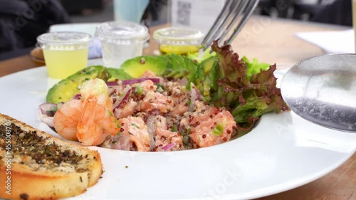 Fresh seafood ceviche mixed with fork and lettuce on side. Chopped salmon, prawns and avocado slices on white plate. Gourmet restaurant food concept